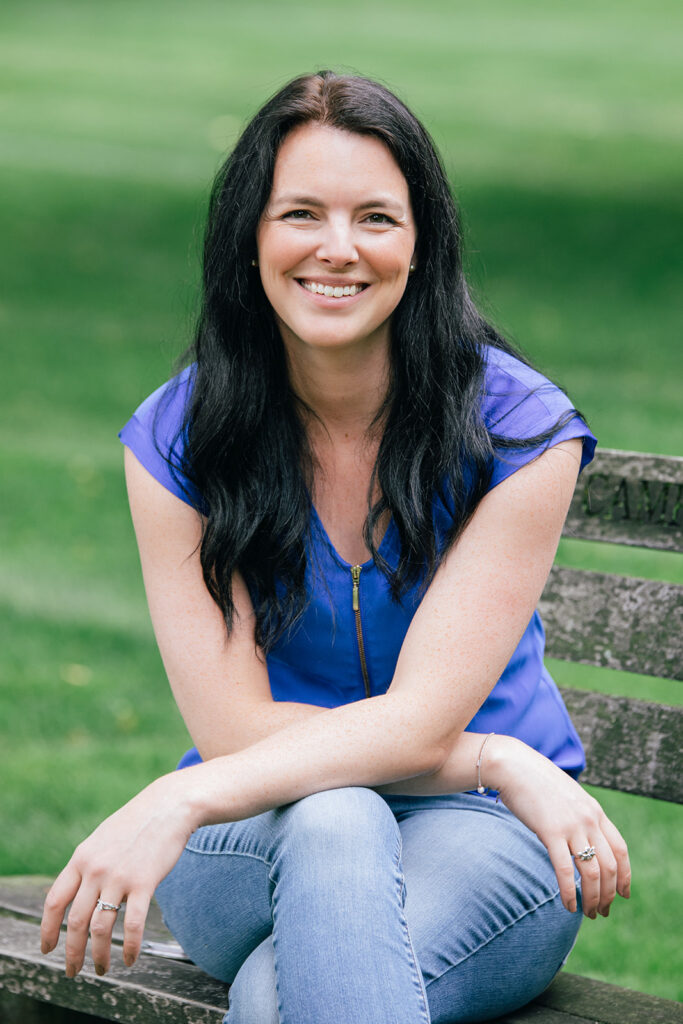 A woman sitting on a bench, leaning forward with her arms crossed, resting on her knees. She's wearing an electric blue shirt, long black hair, and is smiling at the camera.