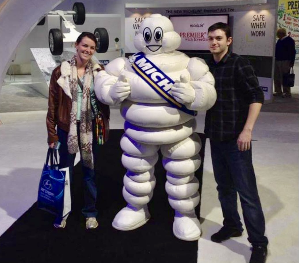 two people standing next to the Michelin man mascot (a large marshmallow looking man with a blue sash that says Michelin), smiling at the camera